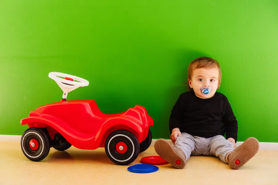 Kleinkind sitzt neben einem roten Bobbycar vor einer grünen Wand